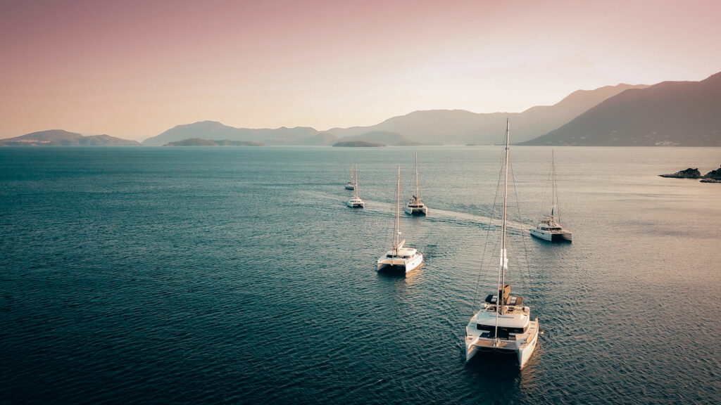 six catamaran boats sailing on the sea with mountains and pink sky in the background